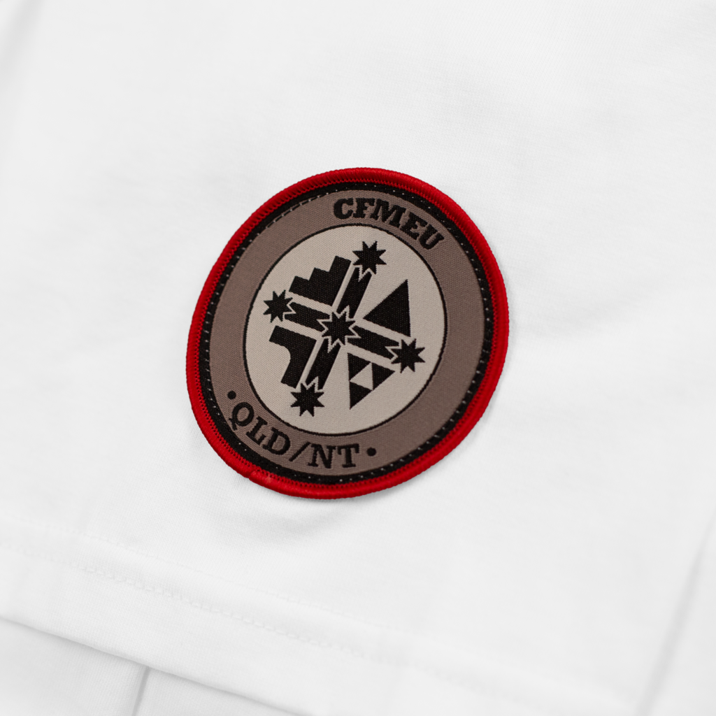 Circular embroidered patch with a red border on white fabric, showing a geometric mountain/star design and partial text around the edge.